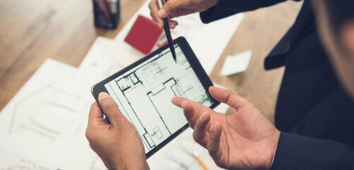 Agente inmobiliario con un cliente o un equipo de arquitectos discutiendo digitalmente, mediante una tableta, un modelo de vivienda y sus planos.